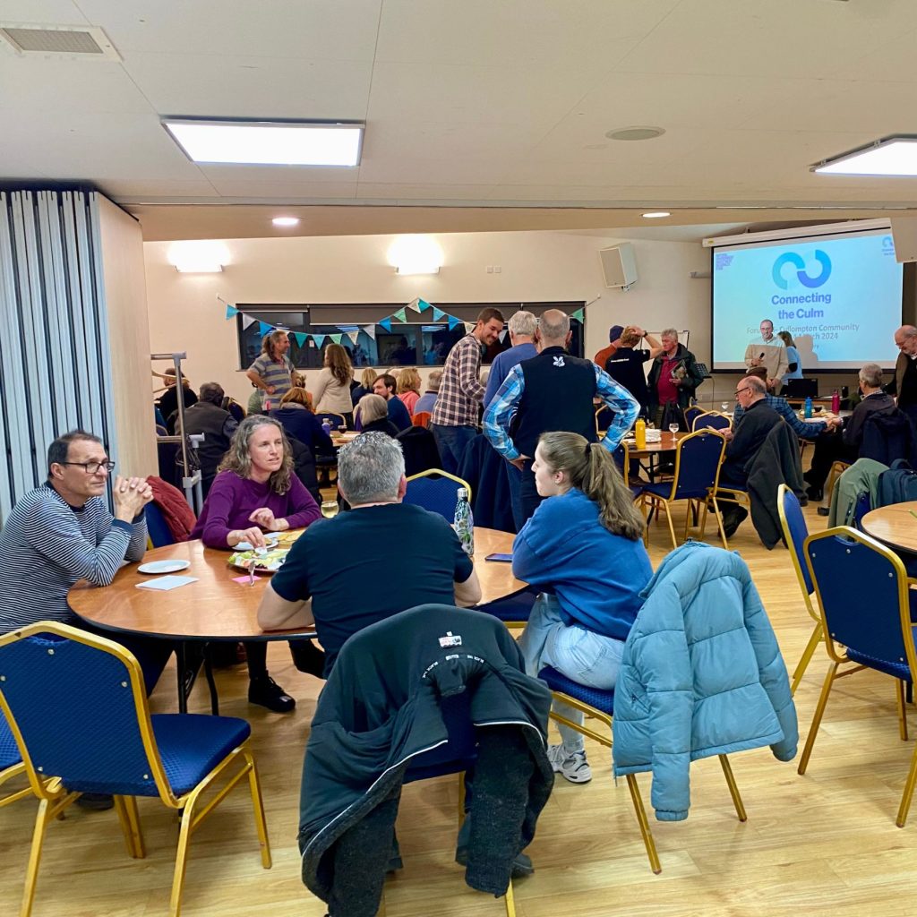 Groups of people have conversation around tables with a presentation screen in the background