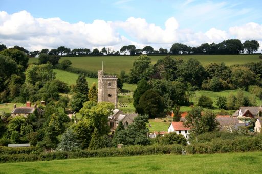 A church tower surrounded by a cluster of houses, fields and trees.